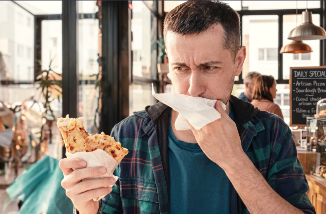 Man covering his mouth looking sick while holding a piece of food.