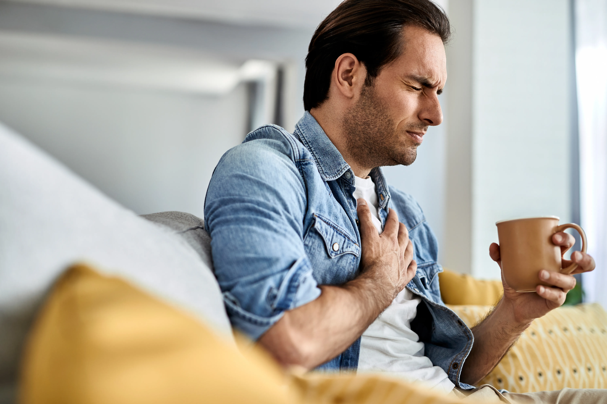 A man sitting on a couch, wincing in pain and holding his chest while holding a mug.