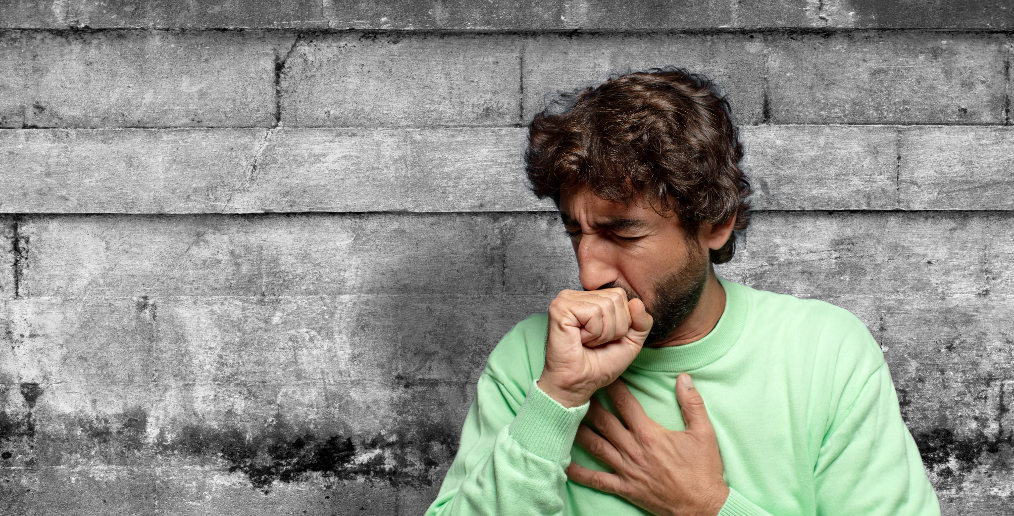 Man in a light green sweater coughing into his fist with a hand on his chest against a gray stone wall