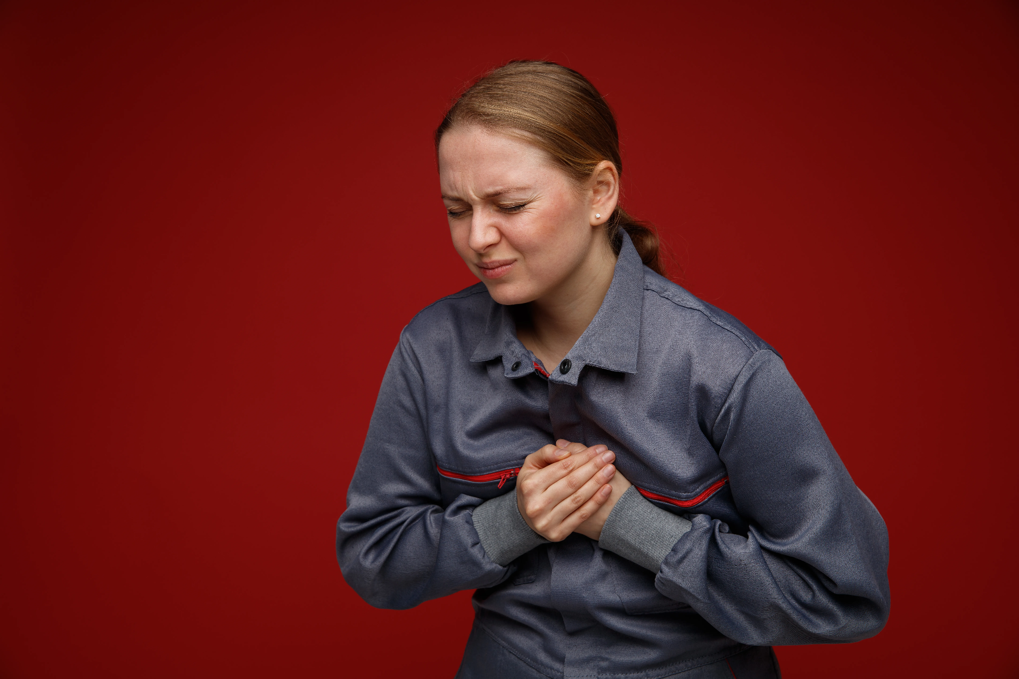 A woman in a grey work uniform holding her chest in pain against a solid red background.