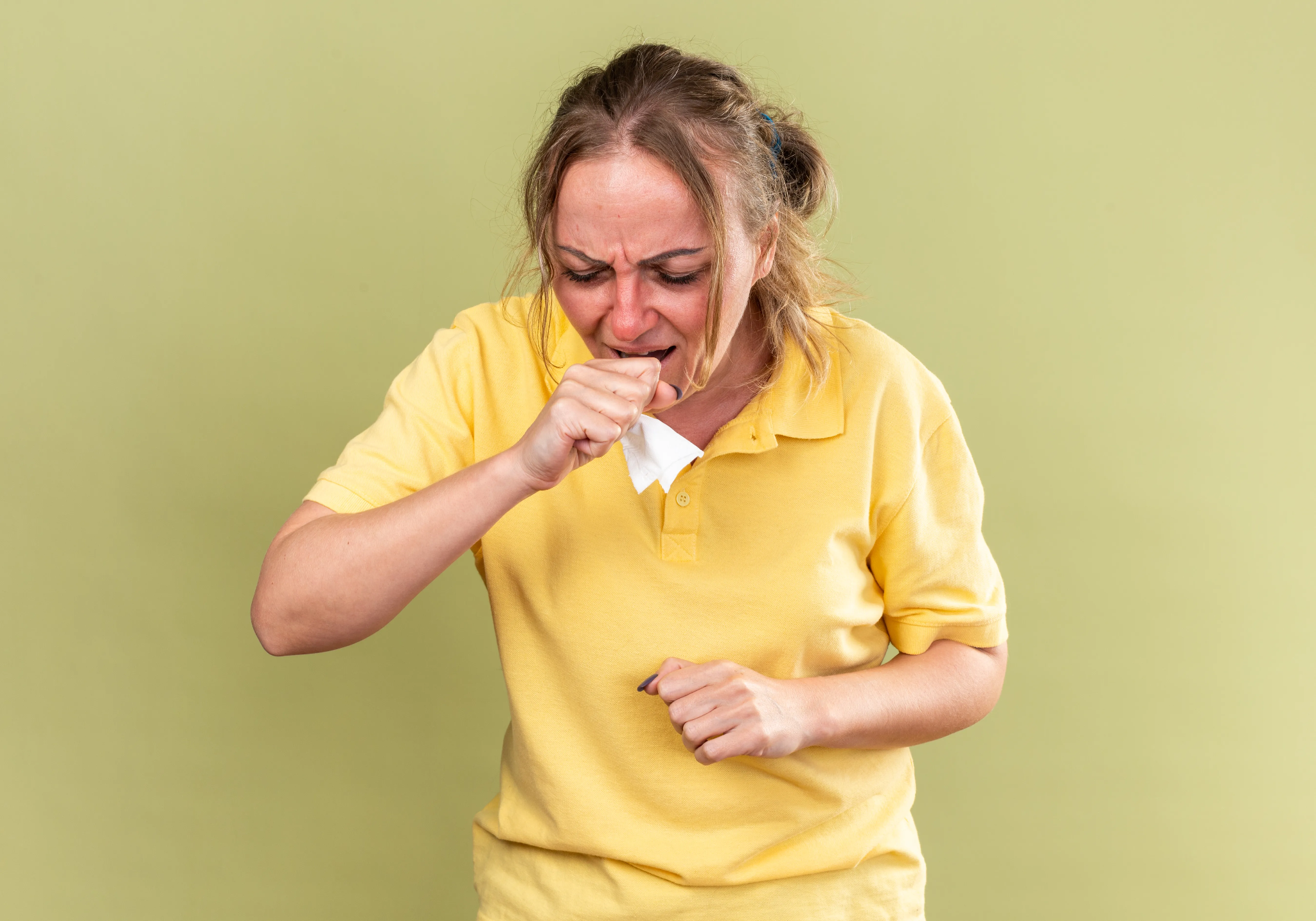 A woman in a yellow polo shirt coughing into a tissue.