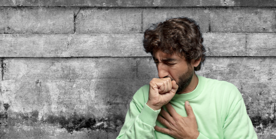 Man in a light green sweater coughing into his fist with a hand on his chest against a gray stone wall
