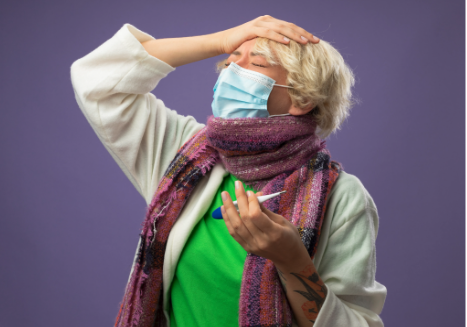 A person wearing a face mask and scarf holding a digital thermometer while touching their forehead in discomfort.