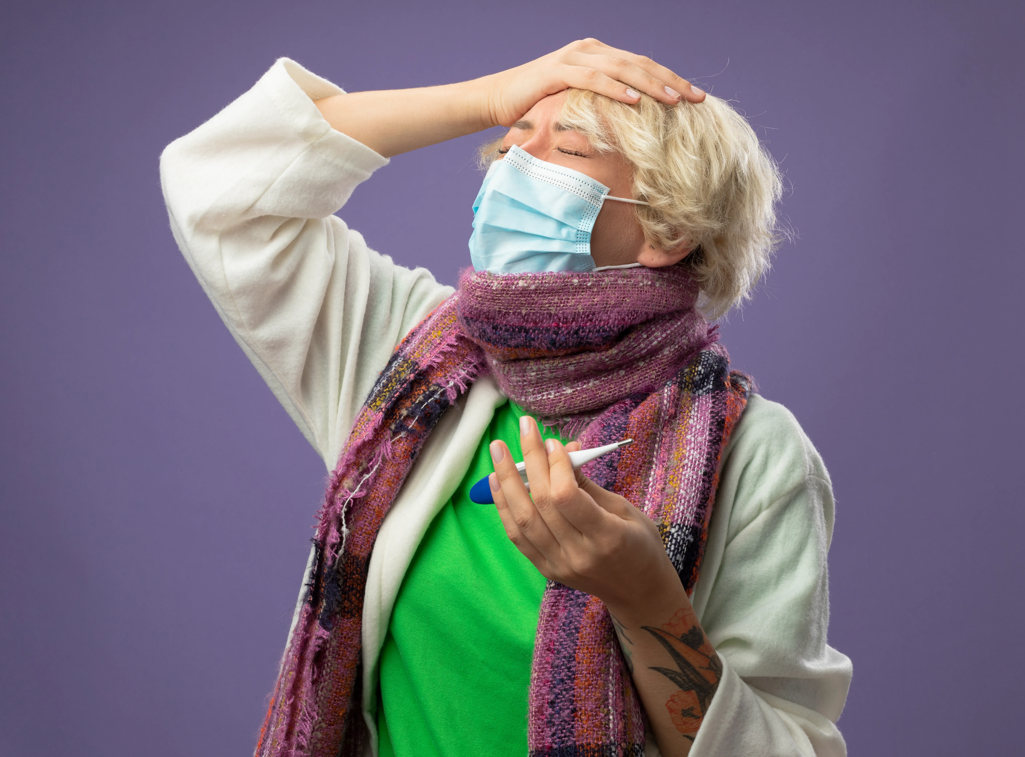 A person wearing a face mask and scarf holding a digital thermometer while touching their forehead in discomfort.