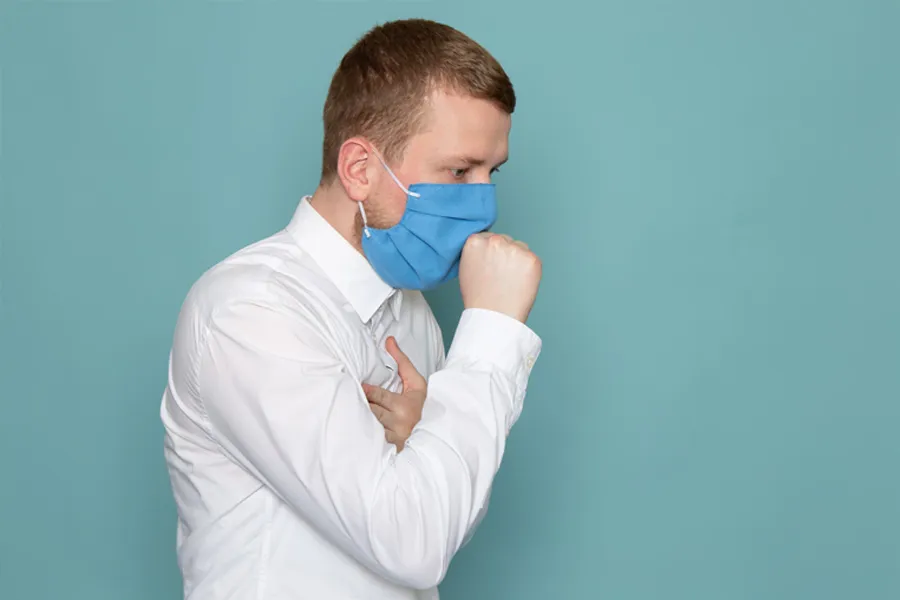 A man wearing a blue surgical mask coughing into his hand while clutching his chest.