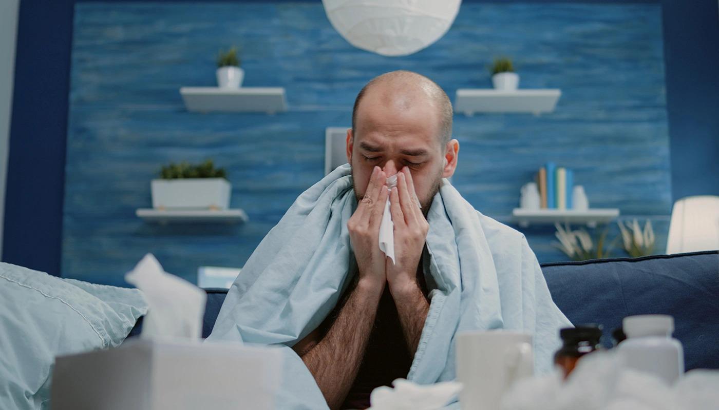 Sick man wrapped in blanket blowing nose with a tissue.
