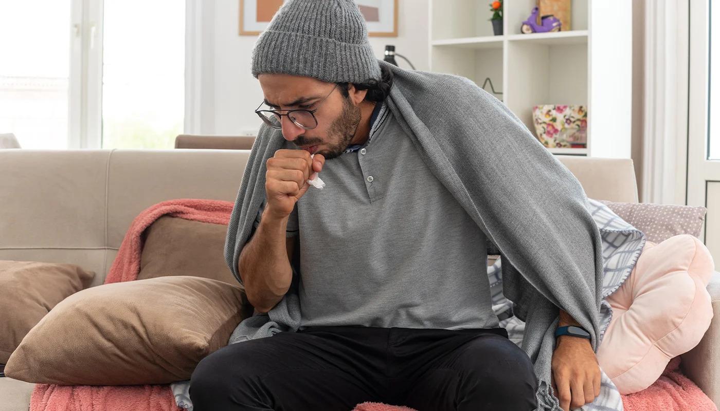 Sick man wrapped in blanket coughing while sitting on sofa
